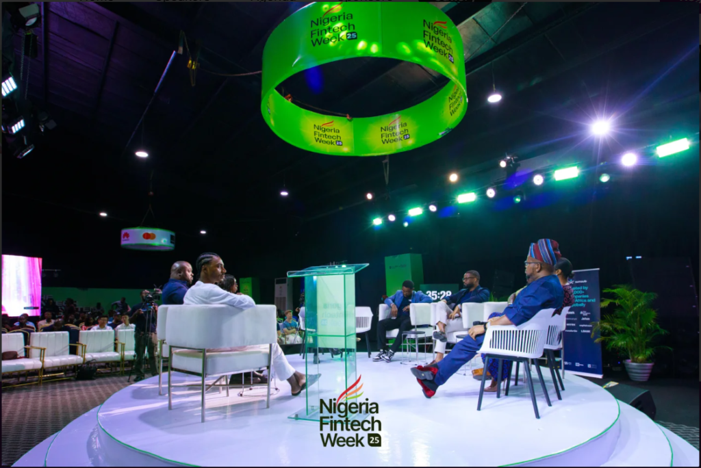 A panel discussion taking place on a circular white stage at an event. A large green circular banner reads 'Nigeria Fintech Week 25' and hangs from the ceiling above a clear podium. Several speakers sit in modern white armchairs facing each other under bright stage lighting, with an audience visible in the background