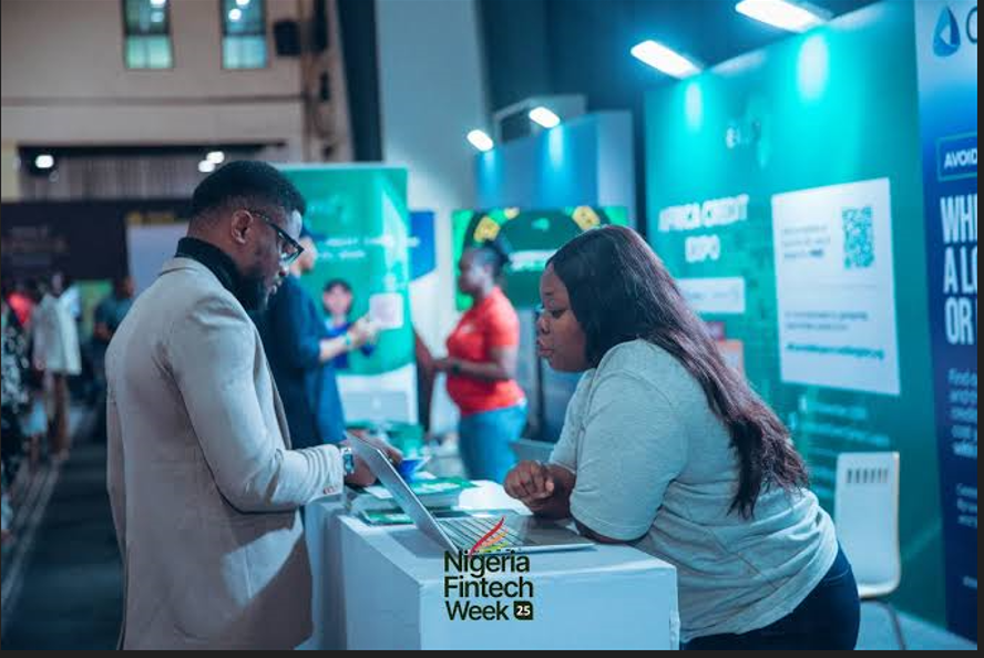 A man in a beige blazer and glasses speaks with a woman at an exhibition counter during Nigeria Fintech Week 25. The woman leans forward over an open laptop computer on the desk, while other attendees and a teal credit expo backdrop are visible in the background.