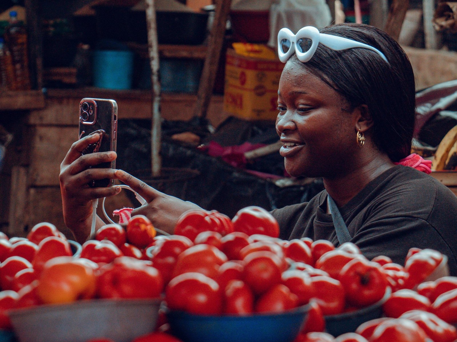 "a dark skinned woman holding up her phone and looking at her it feeling excited. she's in a open stall of her own, and there's a display of tomatoes in little bowls in front her."